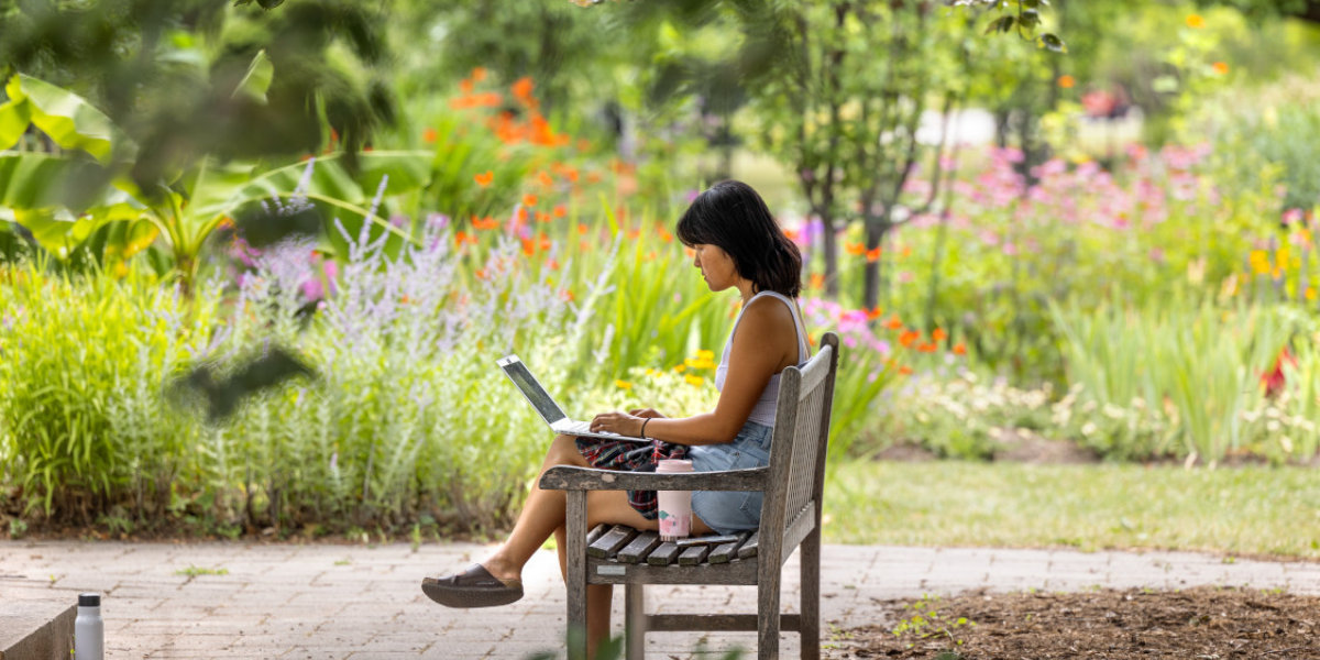 Woman studying on laptop at a outdoor bench, flower garden in the background