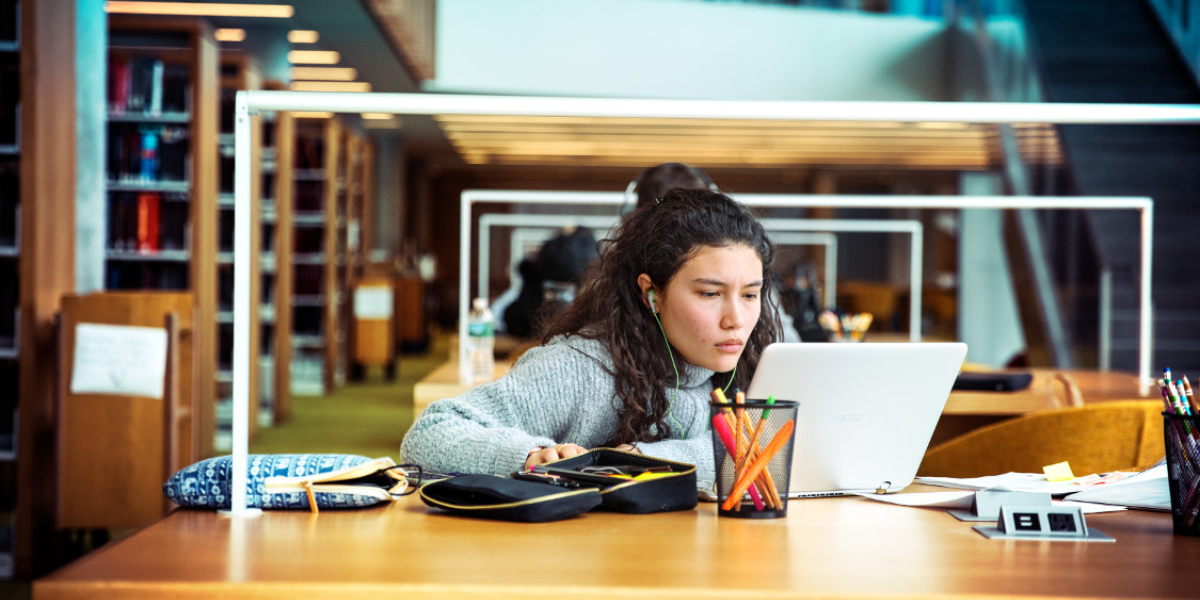 Woman with headphones looking at a laptop at a desk full of study materials