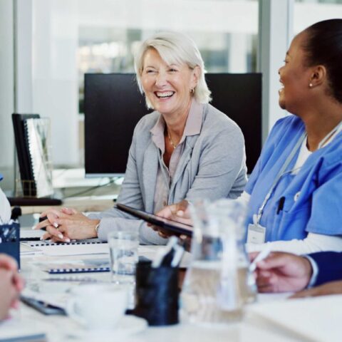 A group of healthcare professionals and a business person sit around a table in a meeting room, smiling and talking during a discussion.
