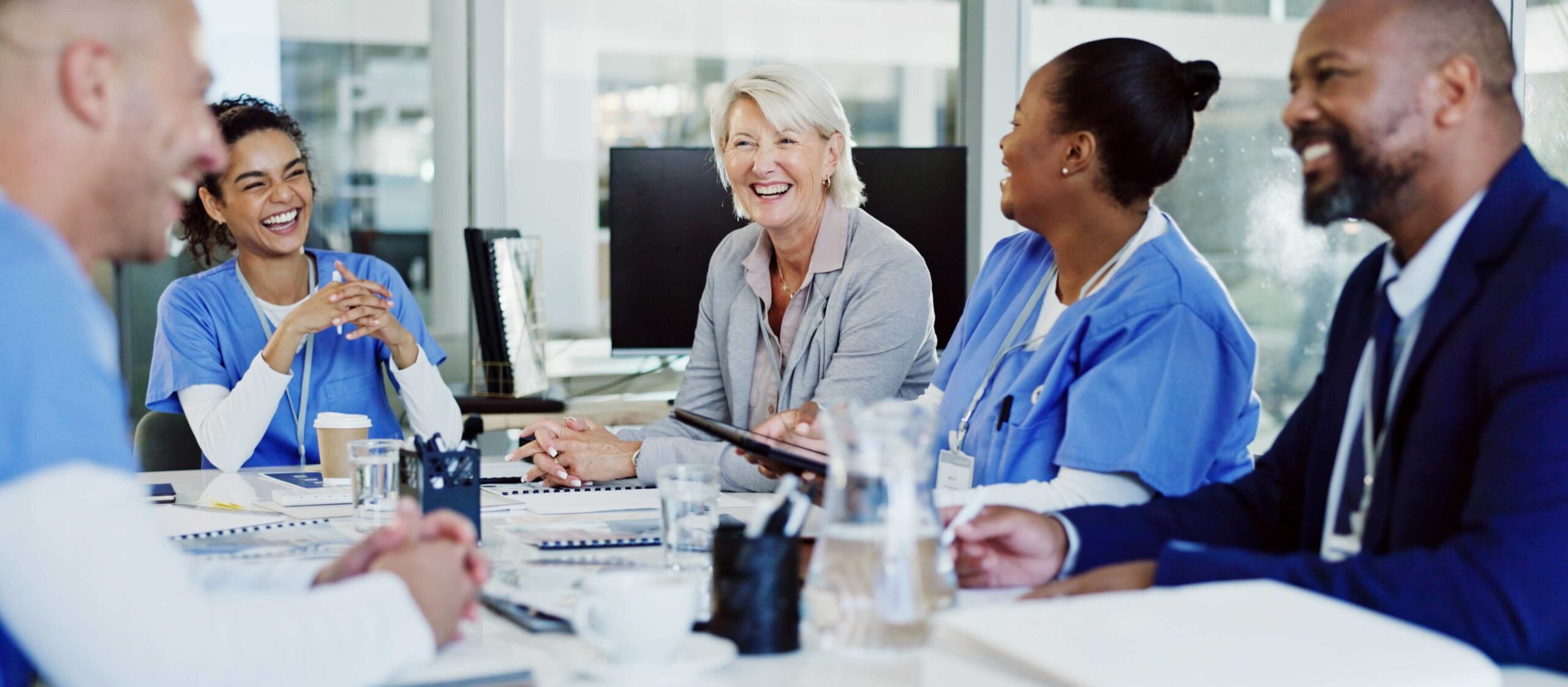 A group of healthcare professionals and a business person sit around a table in a meeting room, smiling and talking during a discussion.