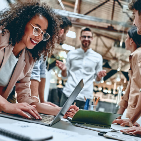 Woman standing and working at computer with a small group of people around a table