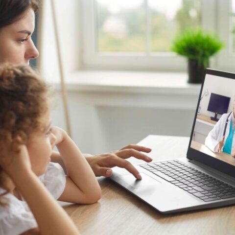 A woman and a young girl sit at a table having a video call with a doctor on a laptop in a bright room.