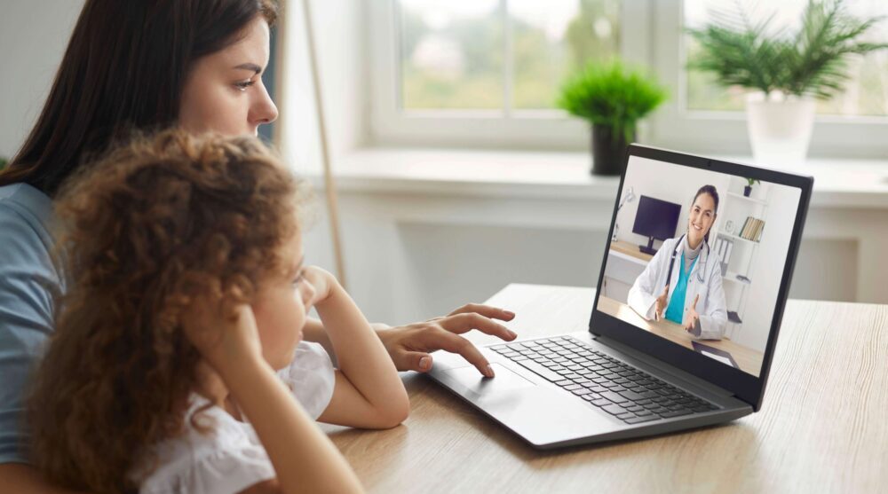 A woman and a young girl sit at a table having a video call with a doctor on a laptop in a bright room.