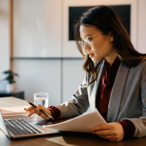 A woman in a blazer sits at a desk, working on a laptop and reviewing documents with a pen in hand. A glass of water and a small plant are on the desk.
