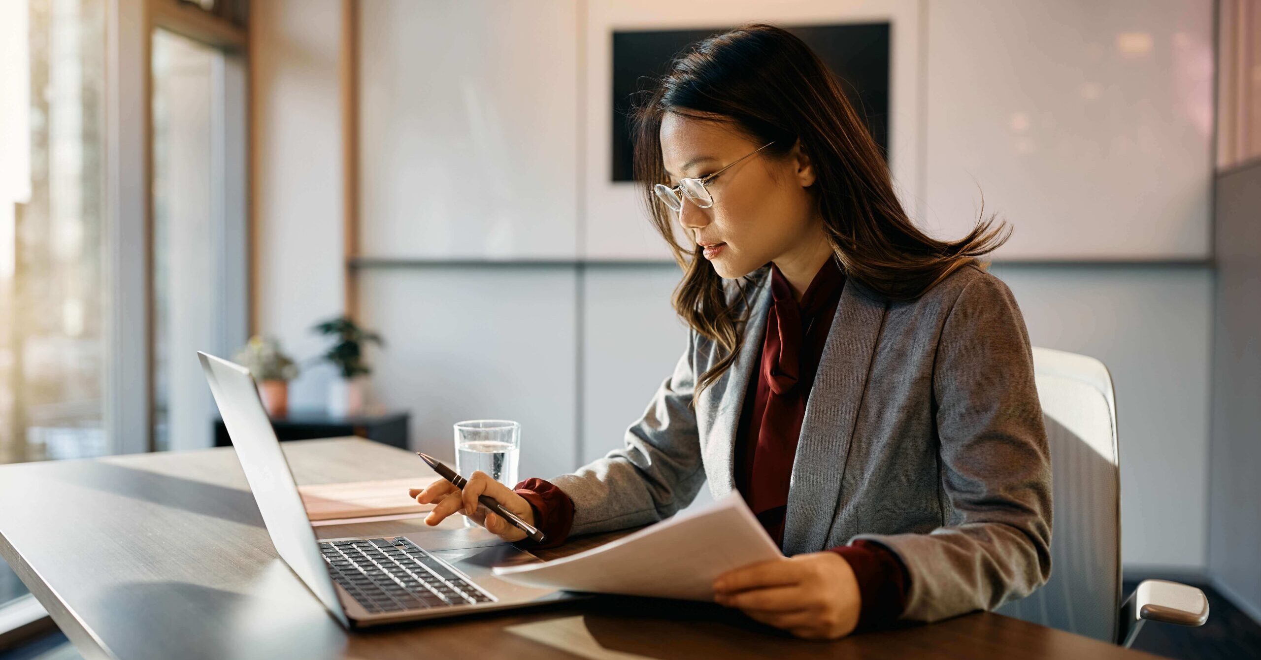 A woman in a blazer sits at a desk, working on a laptop and reviewing documents with a pen in hand. A glass of water and a small plant are on the desk.