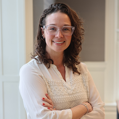 Feminine-presenting person with shoulder length curly brown hair, clear framed eyeglasses, wearing a white long-sleeved blouse, standing and with their arms crossed.